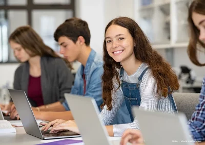Schüler und Schülerinnen sitzen in einem Klassenraum nebeneinander an Ihren Tisch. Vor ihnen steht ein Laptop. Eine junge Schülerin lächelt freundlich in die Kamera.