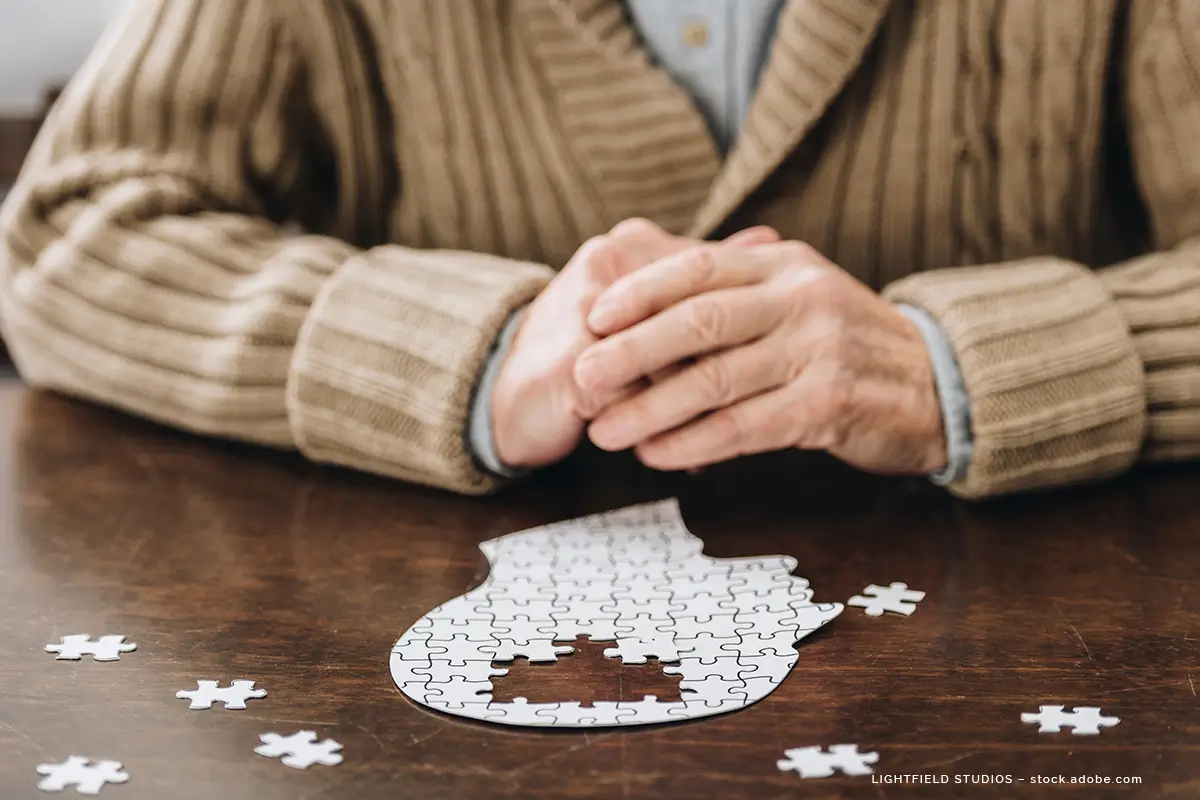 Ein Senior sitzt vor einem Puzzle, welches eine Kopfform darstellt. Im Bereich des Hirns fehlen Puzzleteile.